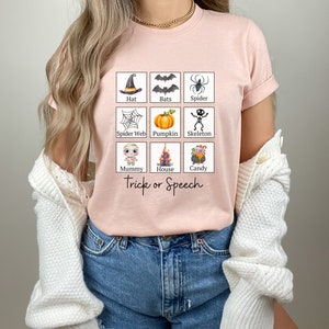 May include: A light pink t-shirt featuring a grid of Halloween-themed illustrations, including a witch's hat, bats, a spider, a spider web, a pumpkin, a skeleton, a mummy, a haunted house, and a cauldron of candy. The words "Trick or Speech" are printed below the grid.