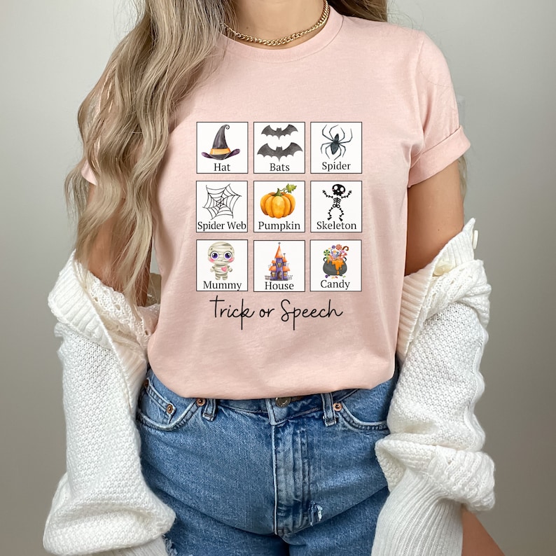 May include: A light pink t-shirt featuring a grid of Halloween-themed illustrations, including a witch's hat, bats, a spider, a spider web, a pumpkin, a skeleton, a mummy, a haunted house, and a cauldron of candy. The words "Trick or Speech" are printed below the grid.