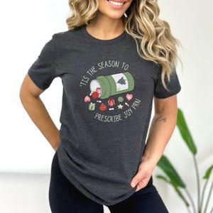 May include: Dark gray t-shirt with a graphic design. The design features a green pill bottle with a Christmas tree and various holiday-themed pills and candies. Text on the shirt reads "'Tis the season to prescribe joy PRN."