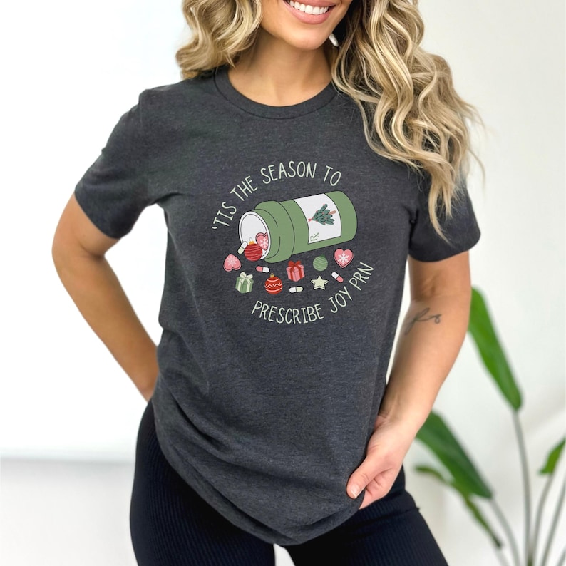 May include: Dark gray t-shirt with a graphic design. The design features a green pill bottle with a Christmas tree and various holiday-themed pills and candies. Text on the shirt reads "'Tis the season to prescribe joy PRN."