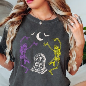 May include: Dark gray t-shirt featuring a Halloween design. The graphic includes two dancing skeletons in purple and yellow, a tombstone with the text "RIP to Gender Roles", a crescent moon, and bats.