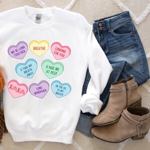 May include: White sweatshirt with colourful heart-shaped graphics, including phrases like "Breathe" and "Lunging for You." The design features hearts in various colours, with text such as "U Had Me At Peep." Shown with blue jeans and tan ankle boots.