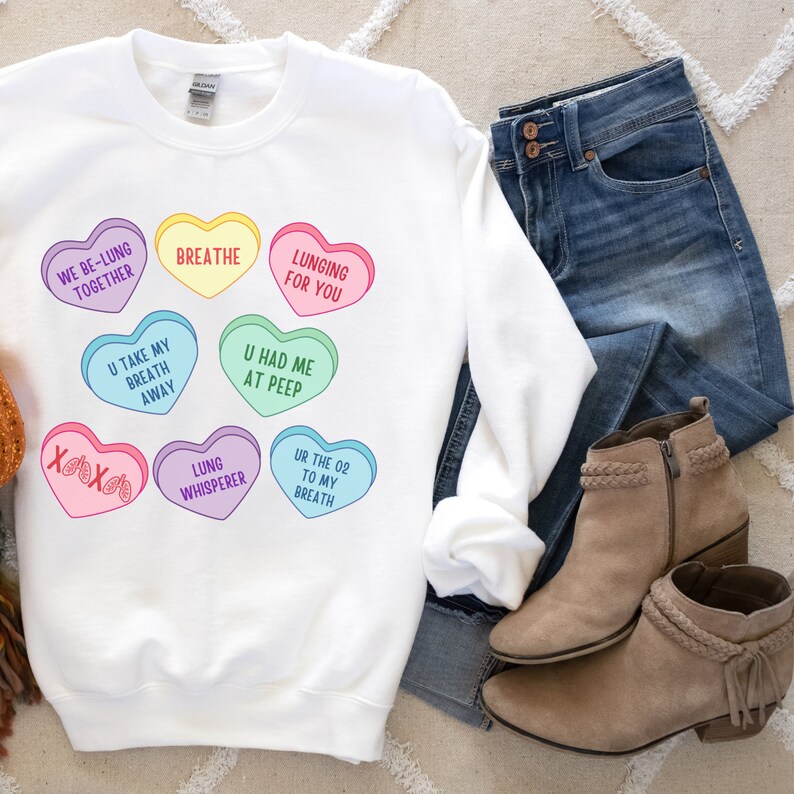 May include: White sweatshirt with colourful heart-shaped graphics, including phrases like "Breathe" and "Lunging for You." The design features hearts in various colours, with text such as "U Had Me At Peep." Shown with blue jeans and tan ankle boots.