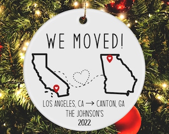 May include: Round white ceramic Christmas ornament with the text "WE MOVED!" and a map of California and Georgia, connected by a dotted line with a heart. The ornament also includes the text "LOS ANGELES, CA -> CANTON, GA THE JOHNSON'S 2022".