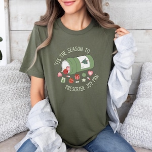 May include: Olive green t-shirt with a graphic design. The design features a green prescription bottle with a Christmas tree and the text "'Tis the season to prescribe joy PRN". Various holiday-themed icons are scattered around the bottle.