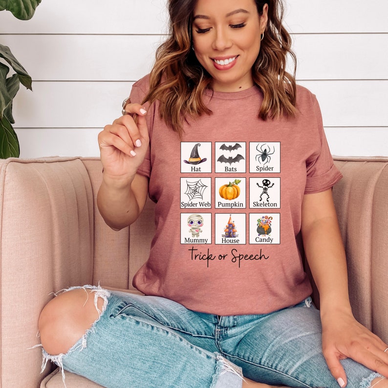 May include: A mauve t-shirt featuring a grid of Halloween-themed illustrations, including a witch's hat, bats, a spider, a spider web, a pumpkin, a skeleton, a mummy, a haunted house, and candy. The text "Trick or Speech" is printed below the grid.