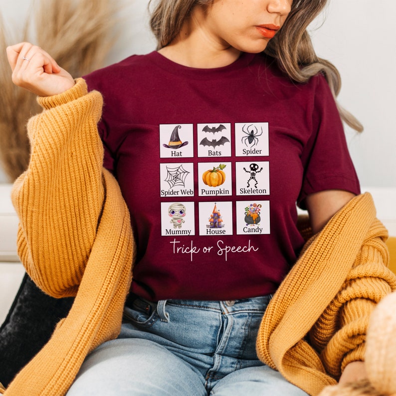 May include: A maroon t-shirt featuring a grid of Halloween-themed illustrations, including a witch's hat, bats, a spider, a spider web, a pumpkin, a skeleton, a mummy, a haunted house, and candy. The text "Trick or Speech" is below the grid.