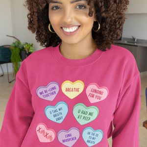 May include: A magenta sweatshirt featuring heart-shaped graphics with phrases like "Breathe" and "Lunging for You." The person wearing the sweatshirt has curly brown hair and gold hoop earrings. The sweatshirt's design includes phrases such as "U Had Me at Peep."