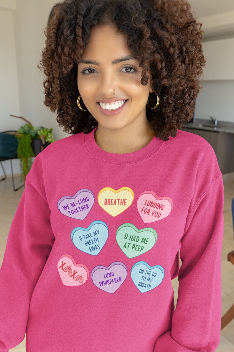 May include: A magenta sweatshirt featuring heart-shaped graphics with phrases like "Breathe" and "Lunging for You." The person wearing the sweatshirt has curly brown hair and gold hoop earrings. The sweatshirt's design includes phrases such as "U Had Me at Peep."