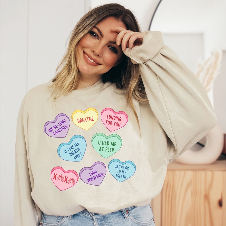May include: A beige sweatshirt with heart-shaped candies displaying phrases such as "Breathe" and "Lunging for You." The design includes phrases such as "We Be-Lung Together" and "U Take My Breath Away." A fun and unique graphic tee.