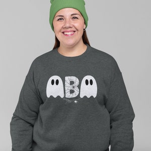 May include: Dark gray sweatshirt with a Halloween-themed design. The design features two white ghost figures flanking the letter "B", with a spider web and spider in the center. The person in the image is wearing a green beanie.