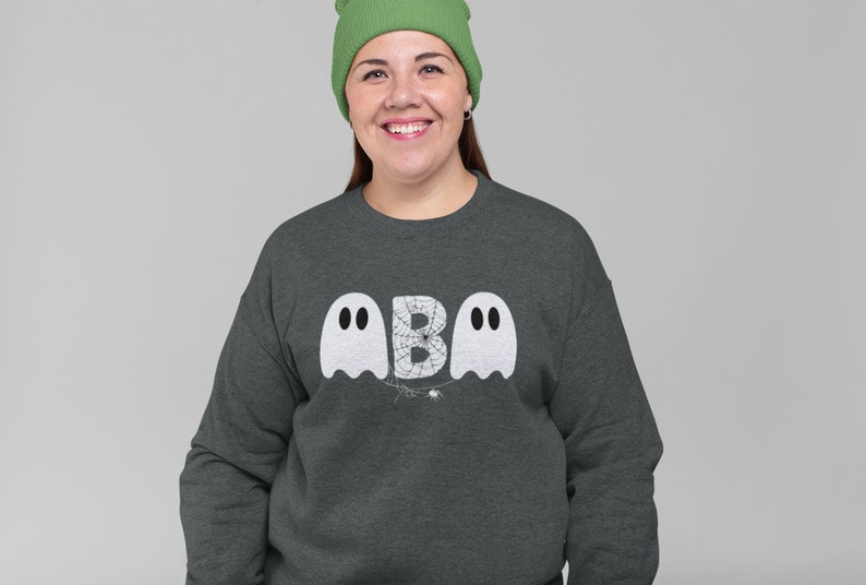 May include: Dark gray sweatshirt with a Halloween-themed design. The design features two white ghost figures flanking the letter "B", with a spider web and spider in the center. The person in the image is wearing a green beanie.