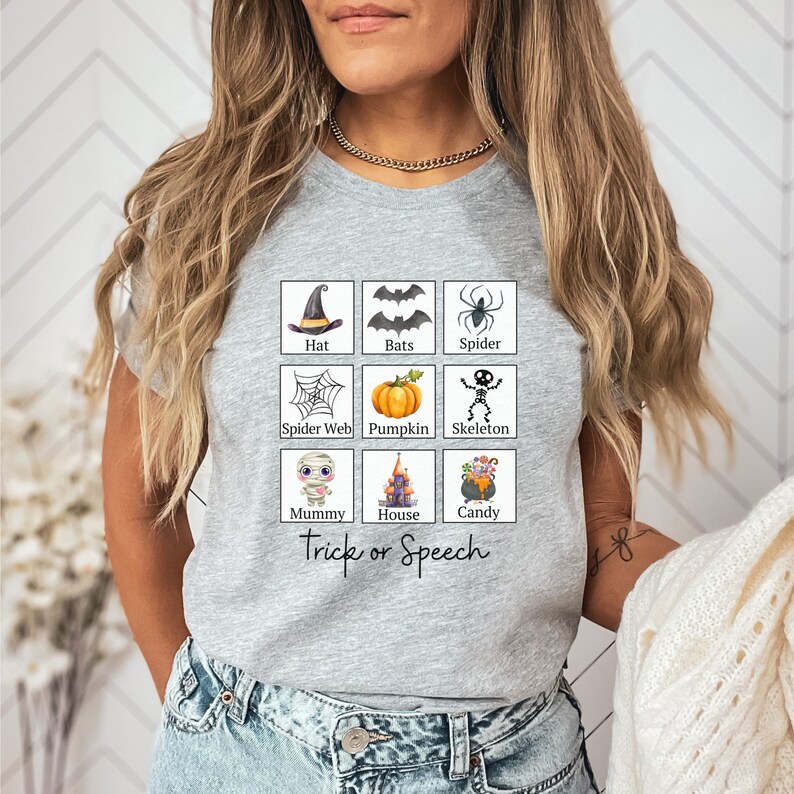 May include: A light gray t-shirt featuring a grid of Halloween-themed illustrations, including a witch's hat, bats, a spider, a spider web, a pumpkin, a skeleton, a mummy, a haunted house, and a cauldron of candy. The text "Trick or Speech" is below the grid.