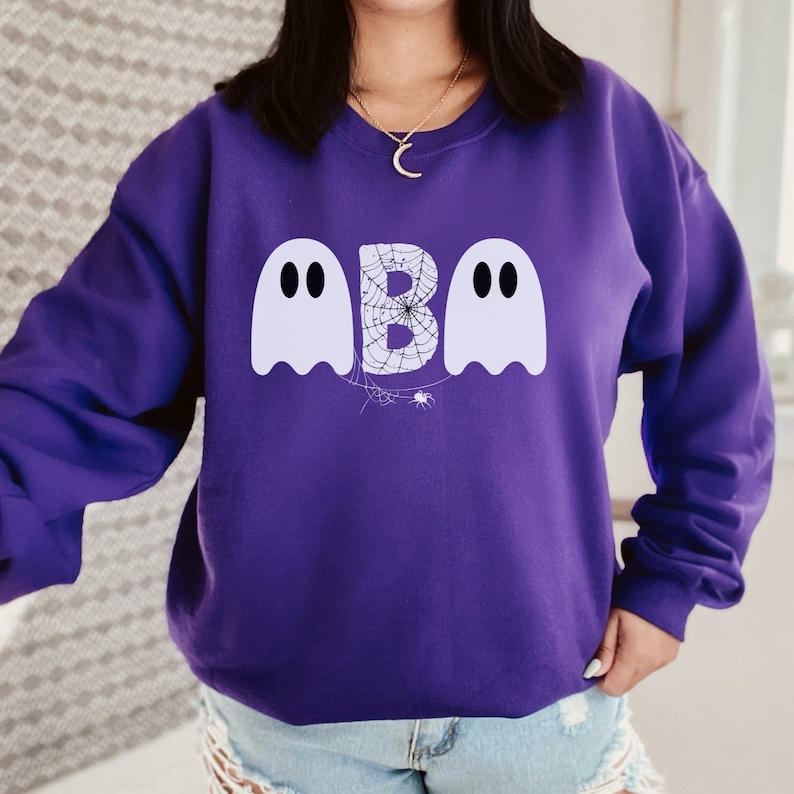 May include: A purple sweatshirt featuring a Halloween-themed design. The design includes two white ghost figures flanking the letter "B", which is filled with a spiderweb pattern. A small spider is also present in the design.