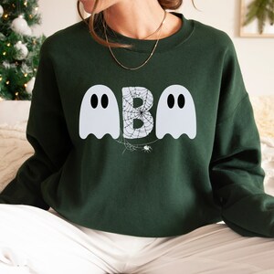 May include: Dark green sweatshirt with a Halloween-themed design. The design features two white ghost figures flanking a letter "B" filled with a spiderweb pattern. A small spider is below the letter "B".