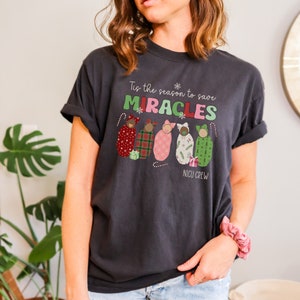 May include: Dark gray t-shirt with the text "Tis the season to save MIRACLES" and "NICU CREW". The shirt features illustrations of swaddled babies in various patterns and colors, with candy canes and gift boxes.