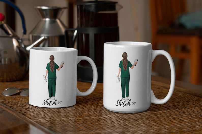Puede incluir: Dos tazas de cer&aacute;mica blanca con una ilustraci&oacute;n verde y blanca de una mujer con bata m&eacute;dica sosteniendo un estetoscopio y una tableta. El texto "Shiloh RT" est&aacute; impreso en la parte inferior de cada taza.