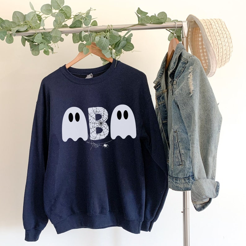 May include: Navy blue sweatshirt with a Halloween ghost design. The design features two white ghost figures flanking a letter "B" with a spiderweb pattern, and a small spider. A denim jacket and straw hat hang nearby.