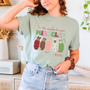 May include: A light green t-shirt with the text "Tis the season to save MIRACLES" and "NICU CREW". The shirt features a graphic of five babies wrapped in patterned blankets. The design includes candy canes and a snowflake.