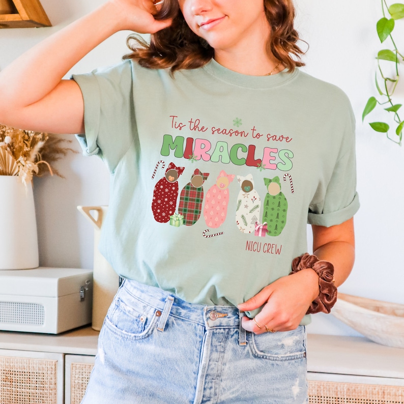 May include: A light green t-shirt with the text "Tis the season to save MIRACLES" and "NICU CREW". The shirt features a graphic of five babies wrapped in patterned blankets. The design includes candy canes and a snowflake.