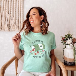 May include: A light green t-shirt with the text "'Tis the Season to Prescribe Joy PRN" and a graphic of a pill bottle with Christmas-themed elements. The shirt is worn by a person.