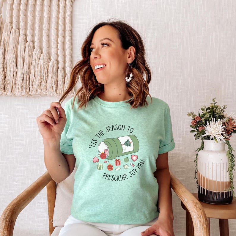 May include: A light green t-shirt with the text "'Tis the Season to Prescribe Joy PRN" and a graphic of a pill bottle with Christmas-themed elements. The shirt is worn by a person.