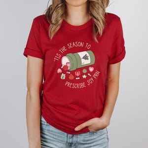 May include: Red t-shirt with the text "'Tis the season to prescribe joy PRN" and a graphic of a green prescription bottle spilling out Christmas-themed pills and decorations. The shirt is a solid red color.