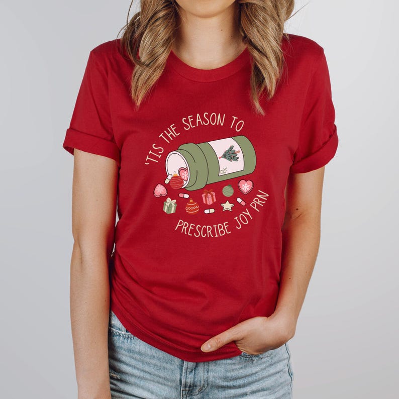 May include: Red t-shirt with the text "'Tis the season to prescribe joy PRN" and a graphic of a green prescription bottle spilling out Christmas-themed pills and decorations. The shirt is a solid red color.