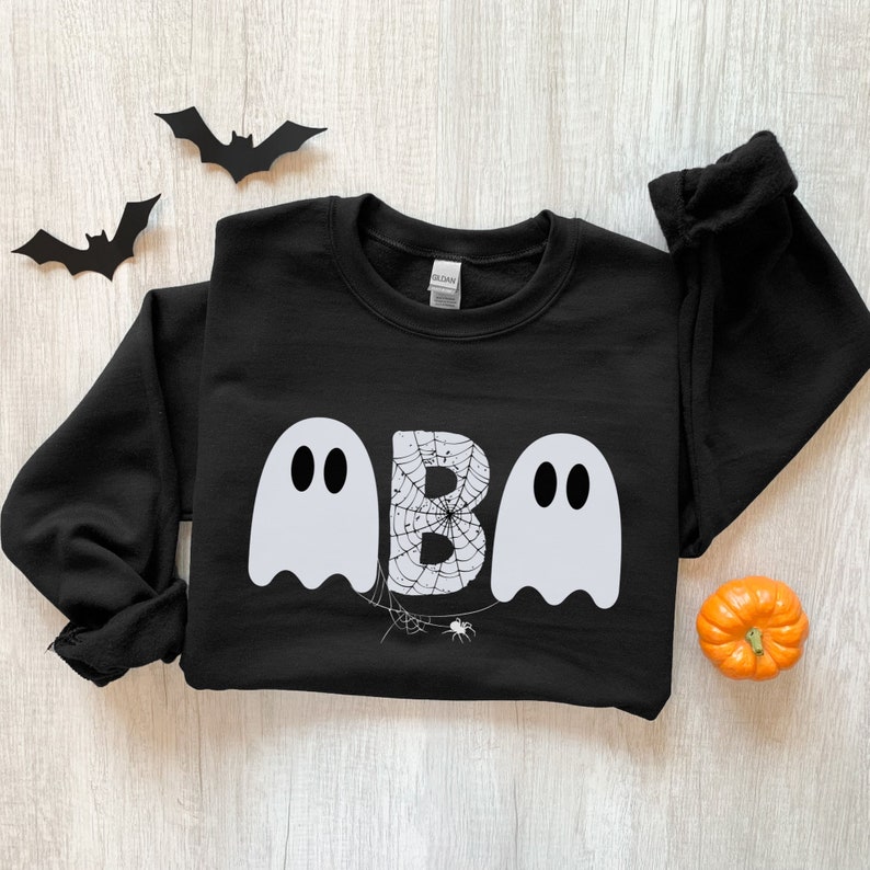May include: Black sweatshirt with a Halloween-themed design. The design features the word "BOO" with the letter "B" in a spiderweb and two ghost figures. Decorative paper bats and a small orange pumpkin are also present.
