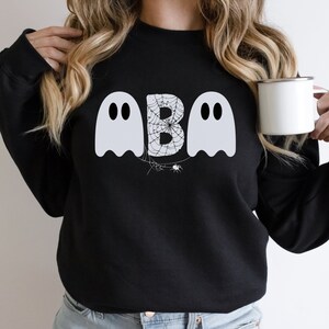 May include: Black sweatshirt featuring a Halloween design. The design includes two white ghost figures flanking a letter "B" filled with a spiderweb pattern. A small spider is below the letter. The sweatshirt is a crew neck style.
