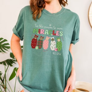 May include: A teal green t-shirt with the text "Tis the season to save MIRACLES" in green, red, and pink. Below the text are illustrations of babies in patterned swaddles. The words "NICU CREW" are printed below the babies.