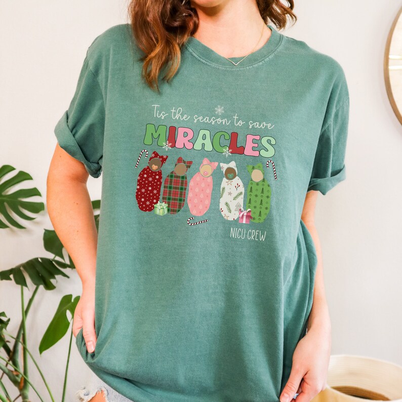May include: A teal green t-shirt with the text "Tis the season to save MIRACLES" in green, red, and pink. Below the text are illustrations of babies in patterned swaddles. The words "NICU CREW" are printed below the babies.