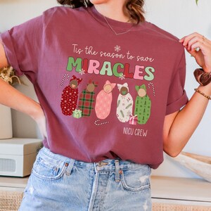 May include: A maroon t-shirt with the text "Tis the season to save MIRACLES" in green, pink, and white. Below the text are illustrations of swaddled babies in various patterns, with candy canes and a gift box. The words "NICU CREW" are at the bottom.