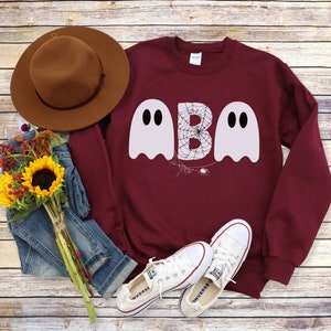 May include: A maroon sweatshirt with the word "BOO" in white ghost-shaped letters. The letter "B" has a spider web design. The image also includes a brown hat, blue jeans, white sneakers, and a bouquet of sunflowers.