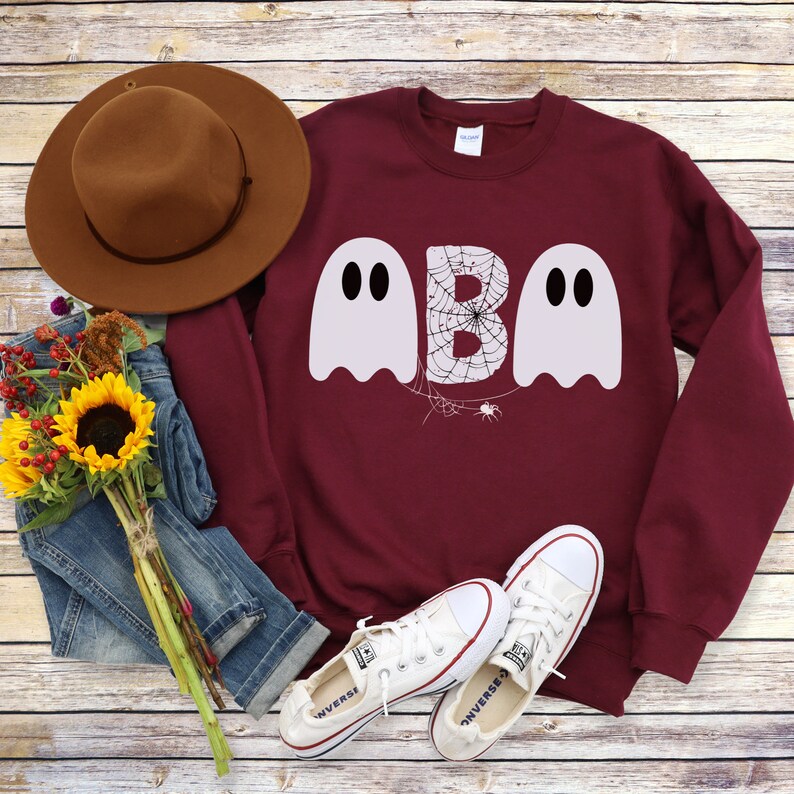 May include: A maroon sweatshirt with the word "BOO" in white ghost-shaped letters. The letter "B" has a spider web design. The image also includes a brown hat, blue jeans, white sneakers, and a bouquet of sunflowers.