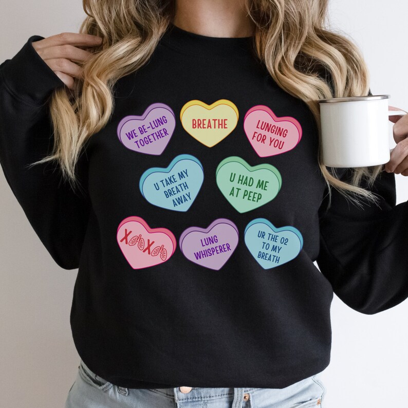 May include: Black sweatshirt with heart-shaped candies displaying phrases such as "Breathe," "Lunging for You," and "We Be-Lung Together." The design uses multiple colours and playful text, creating a fun and unique look.