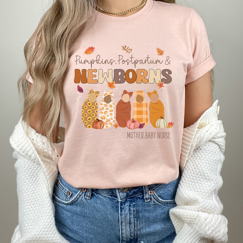 May include: A pale pink t-shirt with the text "Pumpkins, Postpartum & Newborns" in brown and orange. The design includes an illustration of five swaddled babies and pumpkins. The words "Mother Baby Nurse" are printed below the illustration.