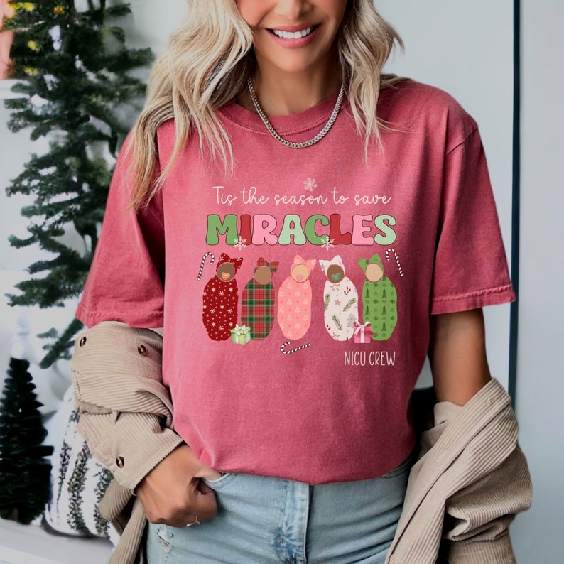 May include: A maroon-colored t-shirt with the text "Tis the season to save MIRACLES" in green and pink. Below the text are five baby swaddles in various patterns and colors. The words "NICU CREW" are at the bottom.