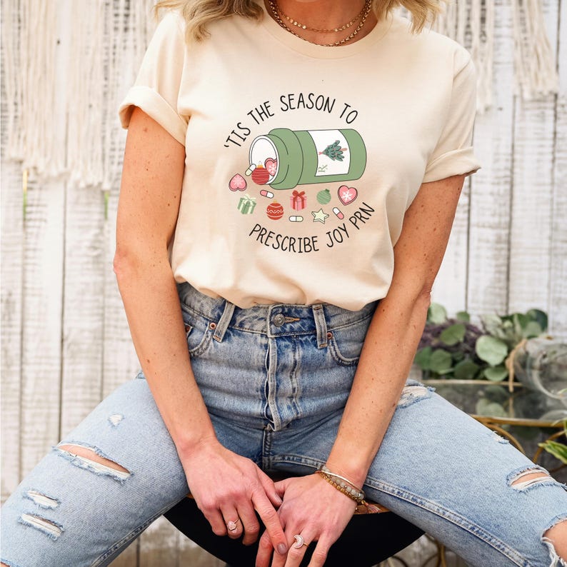 May include: Cream-colored t-shirt with the text "'Tis the Season to Prescribe Joy PRN" and a graphic of a pill bottle spilling out festive pills and ornaments. The shirt is paired with ripped blue jeans.