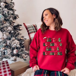 May include: A red sweatshirt with the text "Tis the Season to Shape Behaviors" in green, pink, and white. The sweatshirt has a Santa hat graphic above the text. The person is wearing plaid pants.