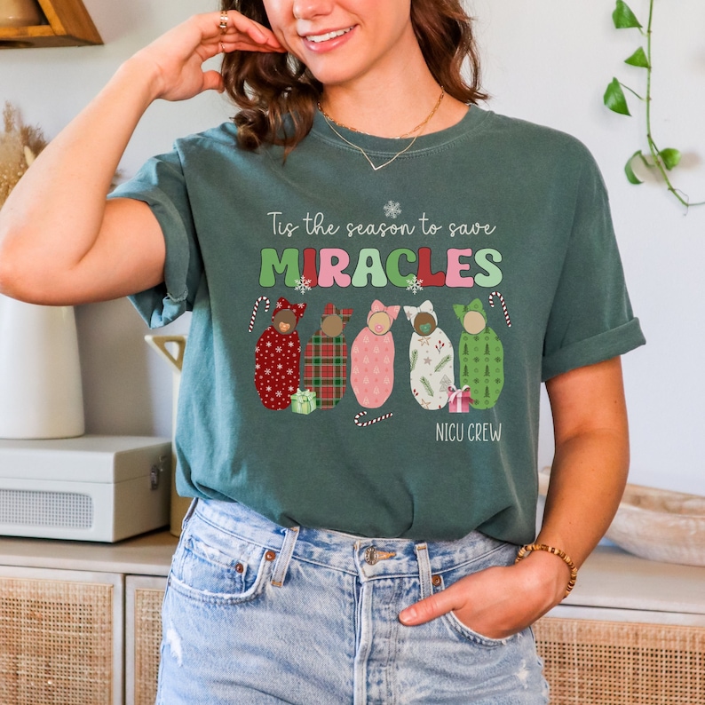 May include: Dark green t-shirt with the text "Tis the season to save MIRACLES" and "NICU CREW". The shirt features illustrations of swaddled babies in festive patterns and candy canes, perfect for the holiday season.