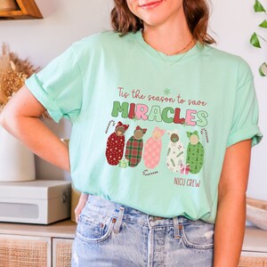 May include: Mint green t-shirt with the text "Tis the season to save MIRACLES" in green, pink, and white. Below the text are five baby illustrations in festive patterns, with candy cane accents. The words "NICU CREW" are at the bottom.