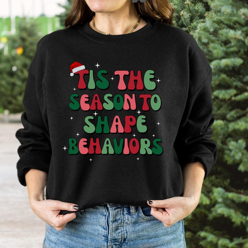 May include: Black sweatshirt with the text "Tis the Season to Shape Behaviors" in red, green, and pink. The design includes a Santa hat and star accents, perfect for the holiday season. A festive and fun apparel item.