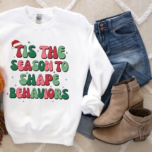 May include: White sweatshirt with the text "TIS THE SEASON TO SHAPE BEHAVIORS" in red and green. The design includes a Santa hat and star accents. The sweatshirt is paired with blue jeans and tan ankle boots.