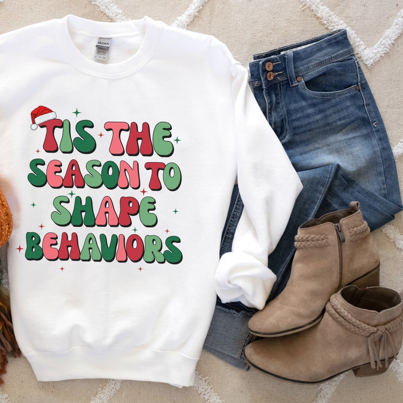 May include: White sweatshirt with the text "TIS THE SEASON TO SHAPE BEHAVIORS" in red and green. The design includes a Santa hat and star accents. The sweatshirt is paired with blue jeans and tan ankle boots.