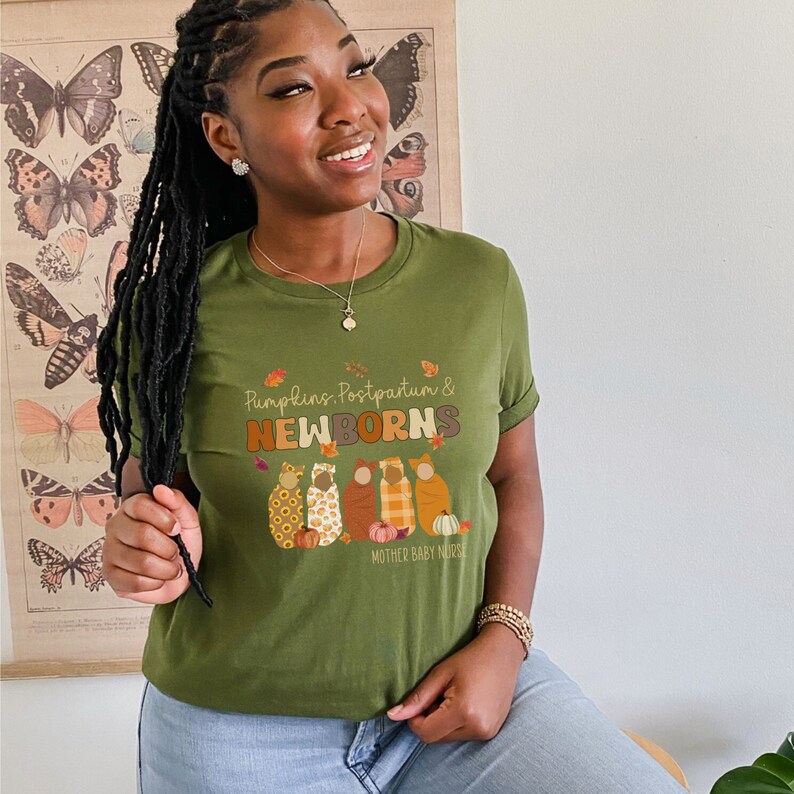 May include: An olive green t-shirt featuring the words "Pumpkins, Postpartum & NEWBORNS" in orange and brown. The design includes four cartoon figures wrapped in blankets, pumpkins, and leaves. The text "MOTHER BABY NURSE" is also present.