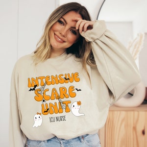 May include: Beige sweatshirt with the text "Intensive Scare Unit" in orange, with bats and spiderwebs. Below the text are two cartoon ghosts. The text "ICU Nurse" is below the ghosts. A Halloween-themed design.