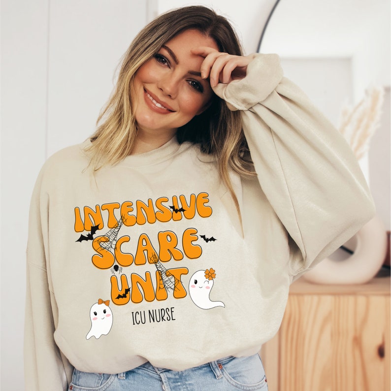 May include: Beige sweatshirt with the text "Intensive Scare Unit" in orange, with bats and spiderwebs. Below the text are two cartoon ghosts. The text "ICU Nurse" is below the ghosts. A Halloween-themed design.