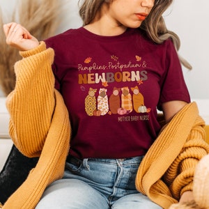 May include: A maroon t-shirt with the words "Pumpkins, Postpartum & NEWBORNS" in a retro font. The shirt features an illustration of five swaddled babies with pumpkins and autumn leaves. The text "MOTHER BABY NURSE" is below the illustration.