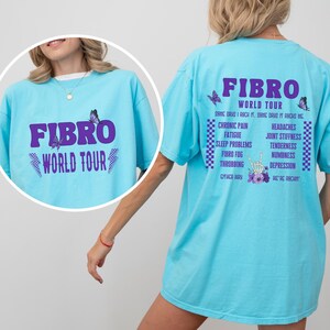 May include: A light blue t-shirt with the text "FIBRO WORLD TOUR" in purple on the front and a list of symptoms associated with fibromyalgia on the back. The back also includes a butterfly graphic and a hand with a peace sign.